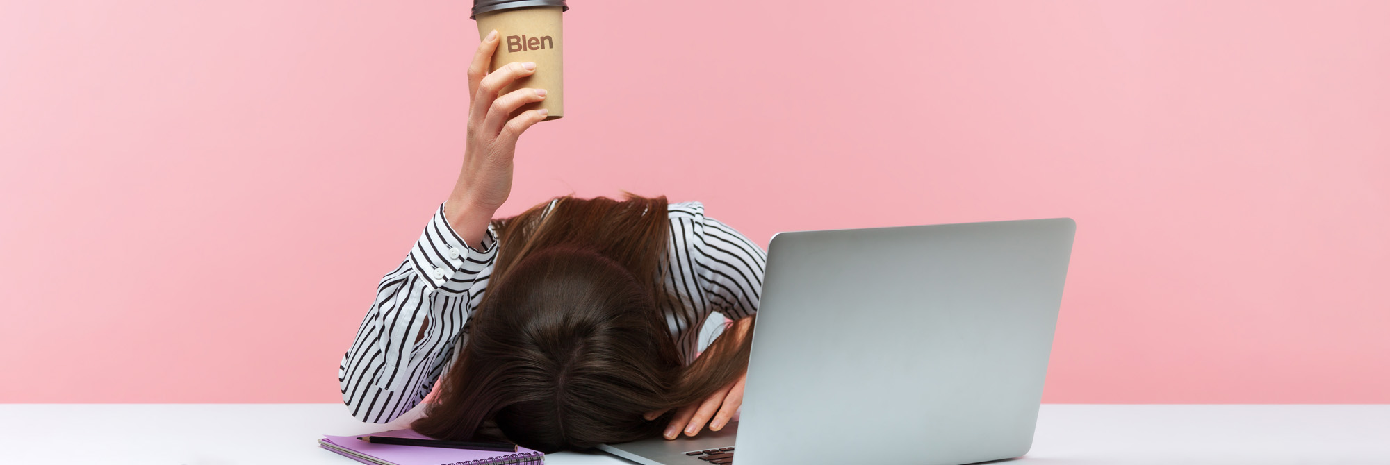 Sleepy woman office worker lying on table with laptop holding and showing paper coffee cup.